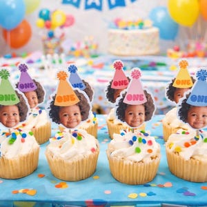 Puede incluir: Cupcakes con glaseado blanco y chispas de colores est&aacute;n decorados con adornos con la cara de un ni&ntilde;o y sombreros de fiesta que dicen "ONE". El fondo incluye globos y un pastel, lo que sugiere una celebraci&oacute;n de primer cumplea&ntilde;os.