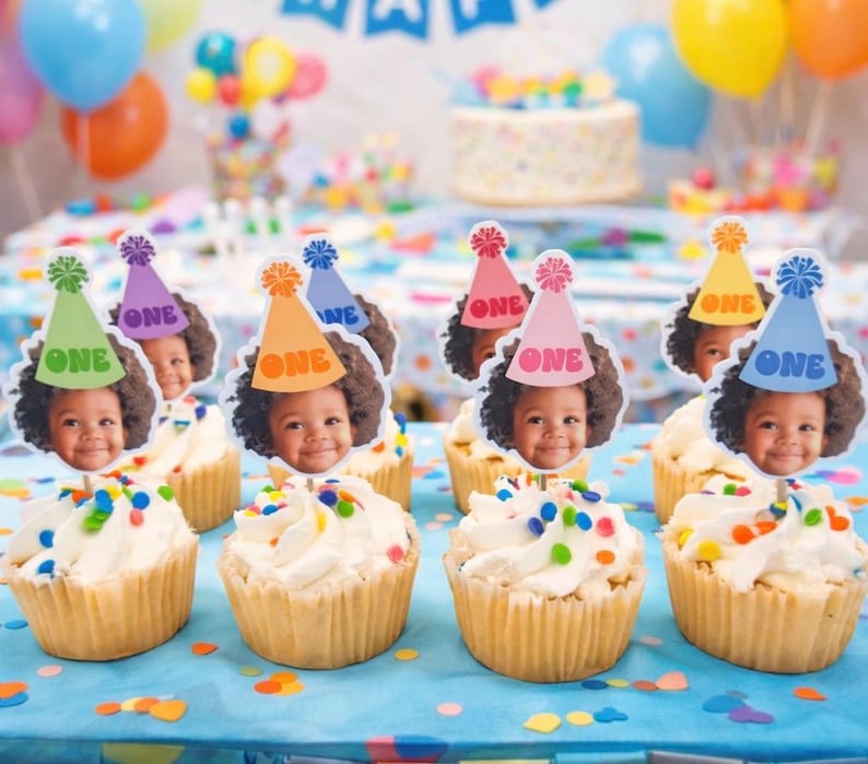 Puede incluir: Cupcakes con glaseado blanco y chispas de colores est&aacute;n decorados con adornos con la cara de un ni&ntilde;o y sombreros de fiesta que dicen "ONE". El fondo incluye globos y un pastel, lo que sugiere una celebraci&oacute;n de primer cumplea&ntilde;os.