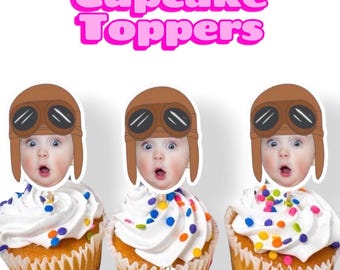 May include: Three cupcakes topped with paper cutouts featuring a baby wearing a brown aviator hat and goggles. The text Cupcake Toppers is displayed in large pink letters above the cupcakes, which are decorated with white frosting and colorful sprinkles.