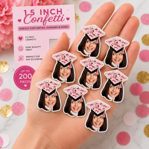 Op de afbeelding: Een verzameling van 3,8 cm grote papieren confetti in de vorm van afstudeerhoeden, elk met een geprint portret van een persoon met donker haar die een hoed draagt versierd met een patroon van roze kersen en strikken, gehouden in een handpalm.