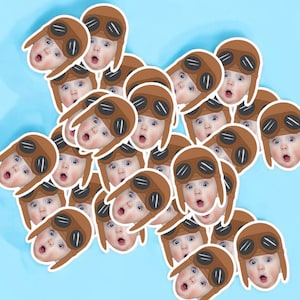 May include: A collection of brown and white stickers featuring a cartoon baby wearing a brown aviator hat and goggles. The stickers are arranged in a random pattern on a light blue background.