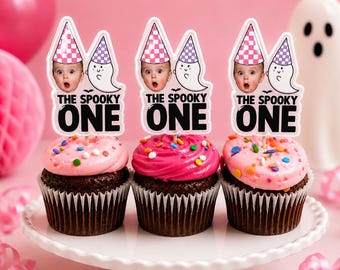 May include: Three chocolate cupcakes with pink frosting and colorful sprinkles, each topped with a paper cake topper featuring a baby's face in a pink checkered party hat, a cartoon ghost in a purple hat, and the text THE SPOOKY ONE.