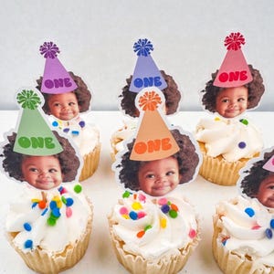 Puede incluir: Cupcakes con glaseado blanco, chispas de colores y adornos con la cara de un ni&ntilde;o y un gorro de fiesta con la palabra "ONE". Los gorros son de varios colores, incluyendo verde, morado, naranja y rosa. Un dulce festivo para un primer cumplea&ntilde;os.