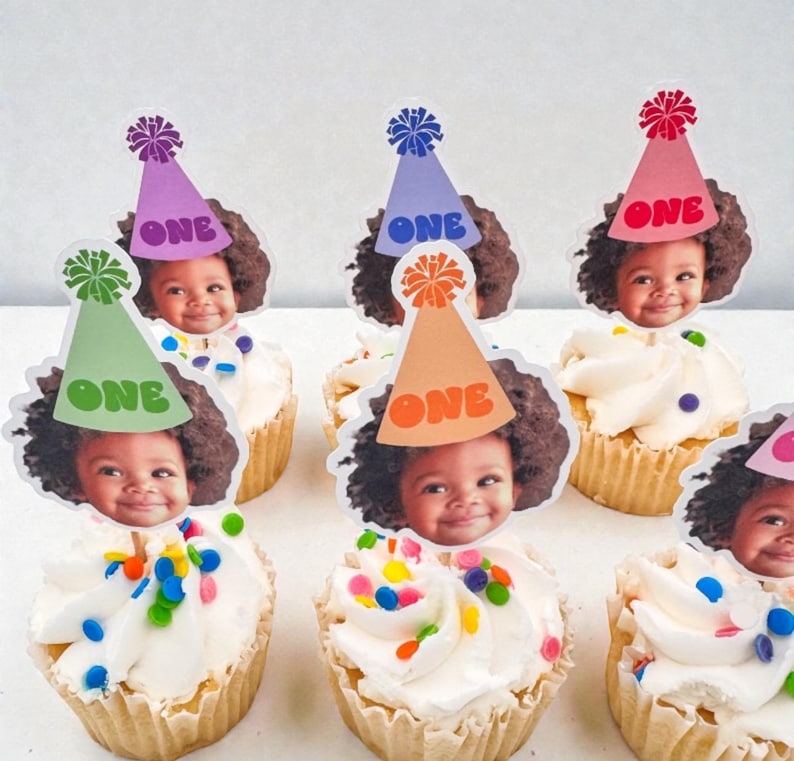 Puede incluir: Cupcakes con glaseado blanco, chispas de colores y adornos con la cara de un ni&ntilde;o y un gorro de fiesta con la palabra "ONE". Los gorros son de varios colores, incluyendo verde, morado, naranja y rosa. Un dulce festivo para un primer cumplea&ntilde;os.