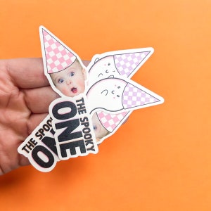 May include: Die-cut stickers featuring a baby's face and a ghost character, both wearing pink and white checkered party hats. The text THE SPOOKY ONE is printed in bold black letters with a pink outline on the bottom of the sticker design.