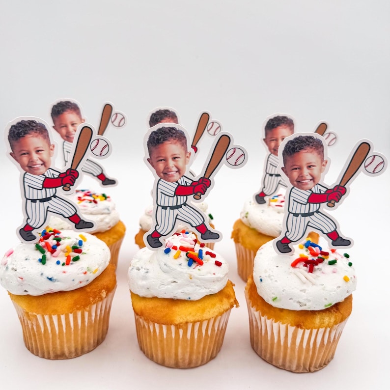 May include: Six cupcakes with white frosting and colorful sprinkles. Each cupcake is topped with a baseball-themed cutout featuring a smiling child holding a bat. The cupcakes have a yellow cake base.