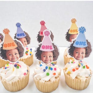 Puede incluir: Cupcakes con glaseado blanco y chispas de colores, cubiertos con im&aacute;genes comestibles de un ni&ntilde;o sonriente con un gorro de fiesta. Cada gorro tiene la palabra "ONE". Los cupcakes est&aacute;n en moldes de papel marr&oacute;n.