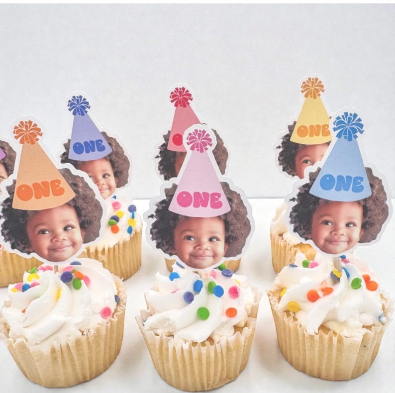 Puede incluir: Cupcakes con glaseado blanco y chispas de colores, cubiertos con im&aacute;genes comestibles de un ni&ntilde;o sonriente con un gorro de fiesta. Cada gorro tiene la palabra "ONE". Los cupcakes est&aacute;n en moldes de papel marr&oacute;n.