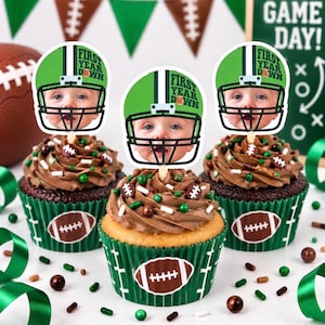May include: Three cupcakes in green wrappers with white football graphics, topped with chocolate frosting and sprinkles. Each cupcake features a paper topper showing a baby's face inside a green football helmet with the text First Year Down.