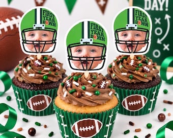 May include: Three cupcakes in green wrappers with white football graphics, topped with chocolate frosting and sprinkles. Each cupcake features a paper topper showing a baby's face inside a green football helmet with the text First Year Down.