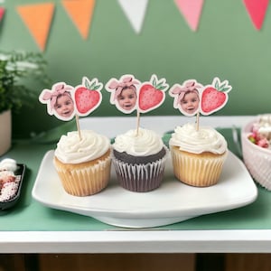 May include: Three cupcakes with white frosting on a white plate. Each cupcake has a decorative topper featuring a child's face and a strawberry. The background includes colorful triangular flags, suggesting a party or celebration.