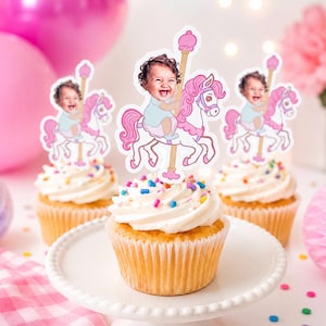 May include: Three cupcakes topped with white frosting and colorful sprinkles, each featuring a paper topper of a smiling baby riding a white carousel horse with a pink mane and tail, attached to a gold-colored pole with a pink top finial.