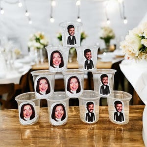 May include: A stack of clear plastic shot glasses, each decorated with a sticker showing a smiling face. The faces alternate between a woman and a man in a tuxedo. The glasses are arranged on a wooden surface.