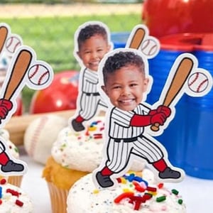May include: Cupcakes with white frosting and colorful sprinkles, topped with baseball-themed cutouts featuring a smiling child in a baseball uniform. Baseball bat and ball decorations are also visible, along with a red helmet and blue cups.