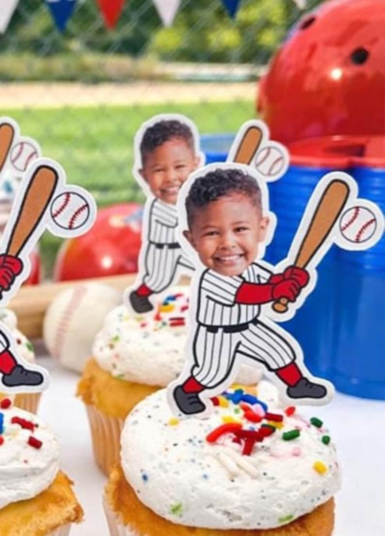 May include: Cupcakes with white frosting and colorful sprinkles, topped with baseball-themed cutouts featuring a smiling child in a baseball uniform. Baseball bat and ball decorations are also visible, along with a red helmet and blue cups.