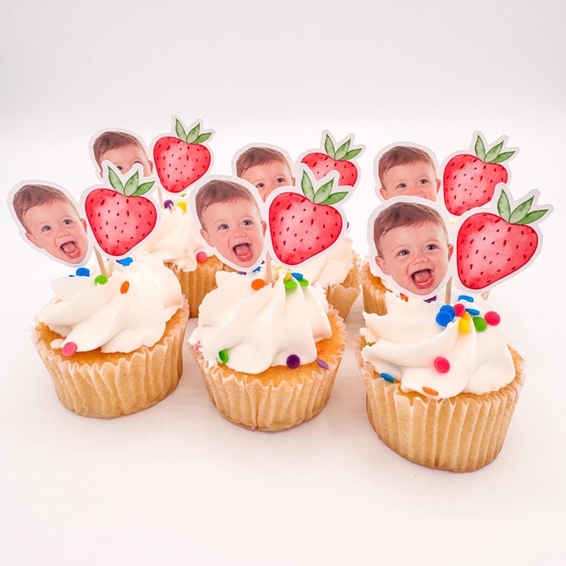 May include: Cupcakes with white frosting and colorful sprinkles, topped with strawberry and baby face toppers. The cupcakes are in a light brown paper cup, perfect for a baby shower or birthday celebration.