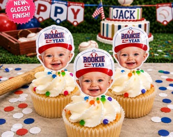May include: Three cupcakes topped with custom photo toppers featuring a smiling baby wearing a red and white baseball cap with the text Rookie of the Year. The cupcakes are decorated with white frosting and colorful sprinkles on a neutral surface.
