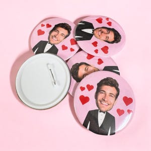 Puede incluir: Cuatro botones redondos de color rosa con la cara de un hombre y un esmoquin en cada uno. Los botones tienen corazones rojos alrededor de la cara del hombre. Un botón se muestra desde la parte posterior, revelando un alfiler plateado.