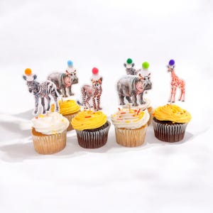 May include: A collection of cupcakes with animal-themed toppers. The cupcakes have yellow and white frosting, and the toppers feature a zebra, hippo, cheetah, and giraffe, each with a colorful pom-pom hat. The cupcakes are arranged on a white surface.