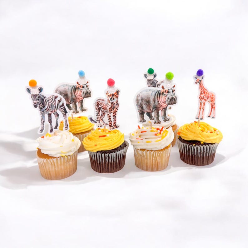 May include: A collection of cupcakes with animal-themed toppers. The cupcakes have yellow and white frosting, and the toppers feature a zebra, hippo, cheetah, and giraffe, each with a colorful pom-pom hat. The cupcakes are arranged on a white surface.