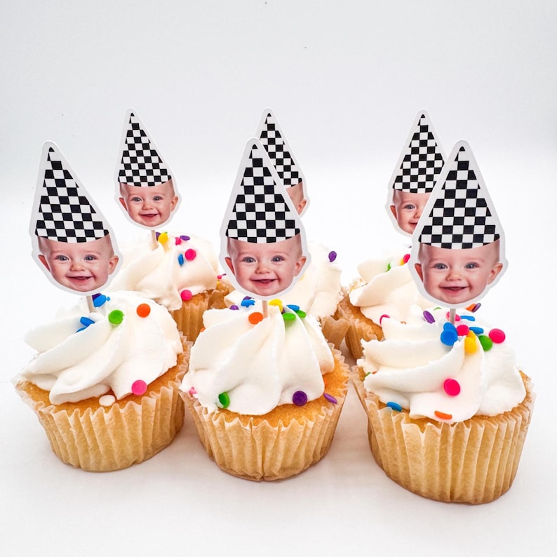 May include: Cupcakes with white frosting and colorful sprinkles, topped with baby face cutouts wearing checkered racing hats. The cupcakes are in paper liners, perfect for a baby shower or birthday party.