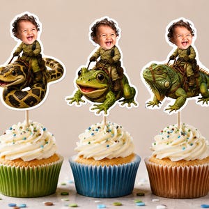 May include: Three cupcakes topped with custom paper toppers featuring a laughing child in a green outfit riding a yellow and black snake, a green frog, and a green iguana. Each topper is mounted on a wooden stick and inserted into white frosting.
