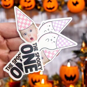 May include: A die-cut sticker featuring a baby's face with a surprised expression wearing a pink and white checkered party hat, alongside a white ghost wearing a matching hat, with the black text THE SPOOKY ONE printed in a bold sans-serif font.