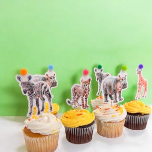 May include: Cupcakes with white and yellow frosting, topped with animal-themed party decorations. The decorations include a zebra, cheetah, hippo, and giraffe, each wearing a party hat with a colorful pom-pom.