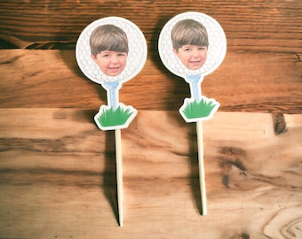 Puede incluir: Dos toppers de cupcake de bola de golf blanca con la cara de un niño en cada bola. Los toppers tienen una base de hierba verde y un palo marrón.