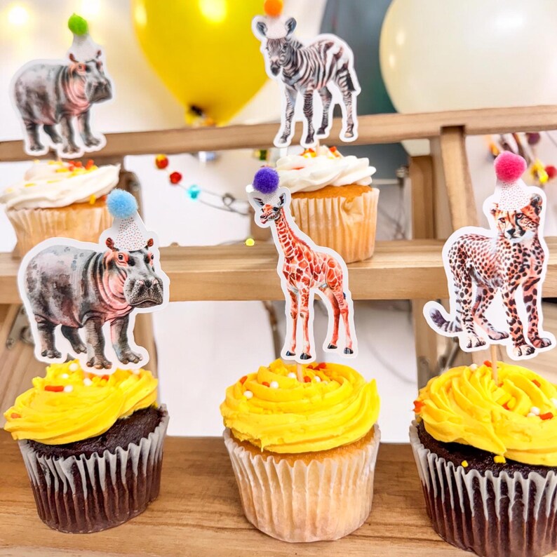 May include: Cupcakes with yellow frosting and animal-themed toppers, including a zebra, giraffe, cheetah, and hippopotamus, each wearing a party hat. The cupcakes are arranged on a wooden tiered stand, with colorful balloons in the background.