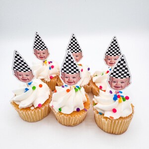 May include: Seven cupcakes with white frosting and colorful sprinkles. Each cupcake is topped with a decorative pick featuring a baby's face wearing a black and white checkered racing flag hat.