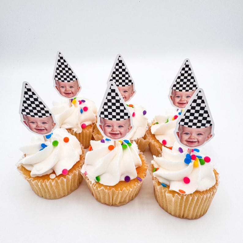 May include: Seven cupcakes with white frosting and colorful sprinkles. Each cupcake is topped with a decorative pick featuring a baby's face wearing a black and white checkered racing flag hat.