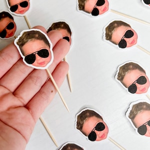 Könnte beinhalten: Eine Sammlung kleiner, runder Aufkleber mit dem Gesicht eines Babys mit schwarzer Sonnenbrille, die an Holzspießen befestigt sind. Die Aufkleber sind weiß mit einem aufgedruckten Bild eines Babygesichts. Die Spieße werden in einer Hand gehalten.