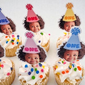 Puede incluir: Cupcakes con glaseado blanco, chispas de colores y una foto de un beb&eacute; con un gorro de fiesta que dice "ONE". Los cupcakes est&aacute;n en un molde para cupcakes marr&oacute;n claro.
