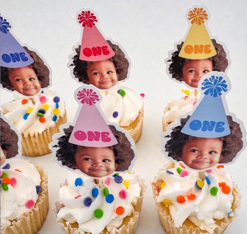 Puede incluir: Cupcakes con glaseado blanco, chispas de colores y una foto de un beb&eacute; con un gorro de fiesta que dice "ONE". Los cupcakes est&aacute;n en un molde para cupcakes marr&oacute;n claro.