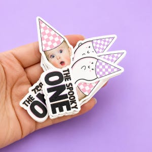 May include: A stack of die-cut stickers featuring a photograph of a baby wearing a pink and white checkered party hat. The text reads THE SPOOKY ONE in bold black letters. Other stickers in the stack show a white ghost wearing a matching party hat.