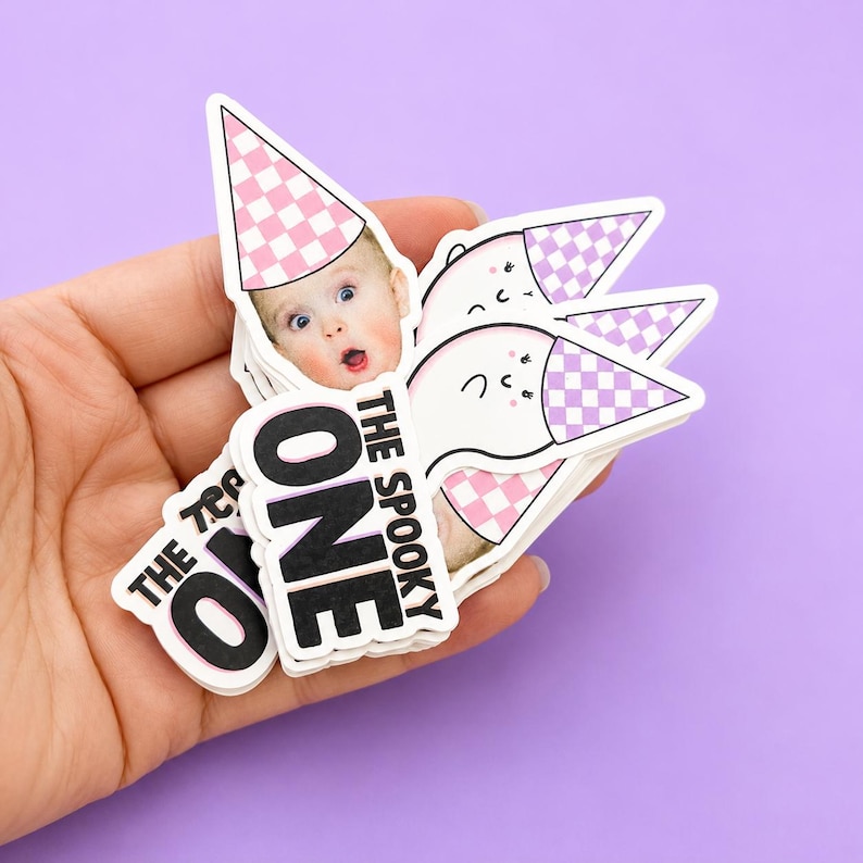 May include: A stack of die-cut stickers featuring a photograph of a baby wearing a pink and white checkered party hat. The text reads THE SPOOKY ONE in bold black letters. Other stickers in the stack show a white ghost wearing a matching party hat.