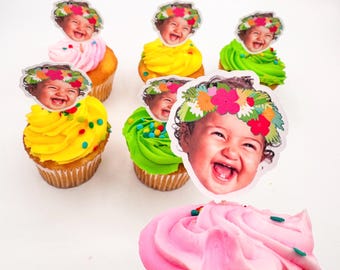 May include: A collection of cupcakes with colorful frosting, including pink, yellow, and green. Each cupcake is topped with a smiling baby face wearing a floral crown. The cupcakes are arranged on a white surface.