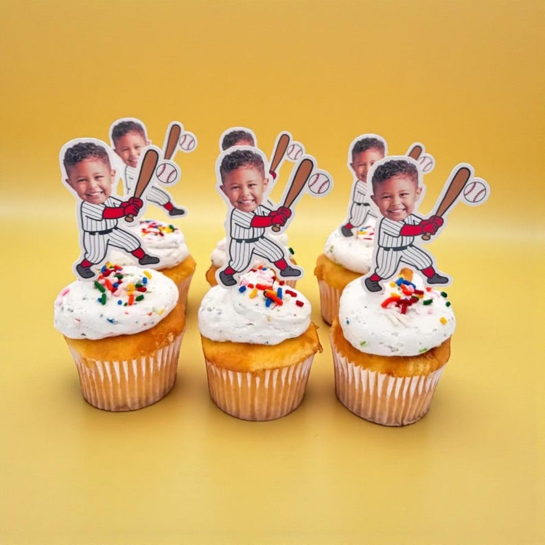 May include: Six cupcakes with white frosting and colorful sprinkles. Each cupcake is topped with a baseball-themed cutout featuring a child holding a bat. The cupcakes sit on a yellow surface.