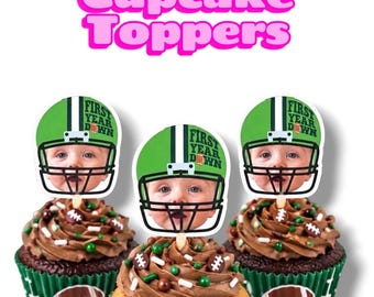 May include: Three cupcakes with chocolate frosting and green football-themed liners featuring a football graphic. Each cupcake has a paper topper showing a baby's face inside a green football helmet with the text First Year Down.
