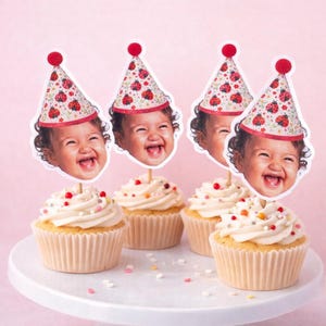 May include: Four cupcakes with white frosting and colorful sprinkles are arranged on a white cake stand. Each cupcake is topped with a photo of a smiling baby wearing a party hat decorated with ladybugs.