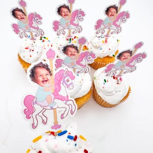 May include: Cupcakes with white frosting and colourful sprinkles. Each cupcake has a carousel-themed topper featuring a smiling baby riding a pink horse. A festive treat for a birthday or celebration.