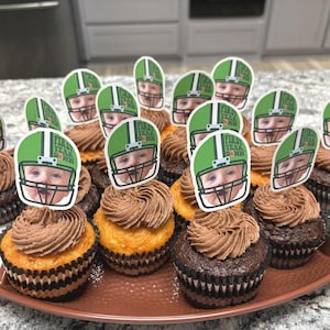 Op de afbeelding: Een verzameling cupcakes met chocolade glazuur en decoratieve toppers. De toppers tonen het gezicht van een baby in een groene voetbalhelm met de tekst "First Year Down". De cupcakes zijn gerangschikt op een bruine schaal.