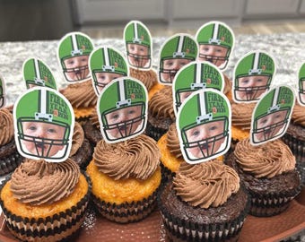 Può includere: Una collezione di cupcakes con glassa al cioccolato e decorazioni. Le decorazioni presentano il volto di un bambino all'interno di un casco da football verde con la scritta "First Year Down". I cupcakes sono disposti su un vassoio marrone.