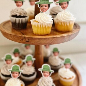 Op de afbeelding: Een houten etag&egrave;re met cupcakes met witte frosting. Elke cupcake is versierd met een decoratieve prikker met het gezicht van een kind met een groene hoed met de tekst "HAPPY CAMPER". De cupcakes hebben gele en chocolade bodems.