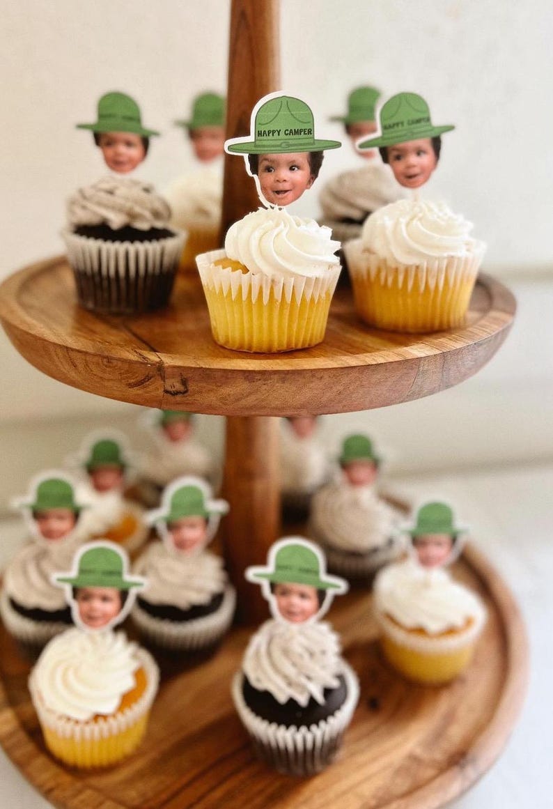 Op de afbeelding: Een houten etag&egrave;re met cupcakes met witte frosting. Elke cupcake is versierd met een decoratieve prikker met het gezicht van een kind met een groene hoed met de tekst "HAPPY CAMPER". De cupcakes hebben gele en chocolade bodems.