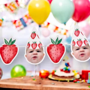 May include: A white banner with red and green strawberries and baby faces. The banner is strung with white string and has a party hat on each strawberry.