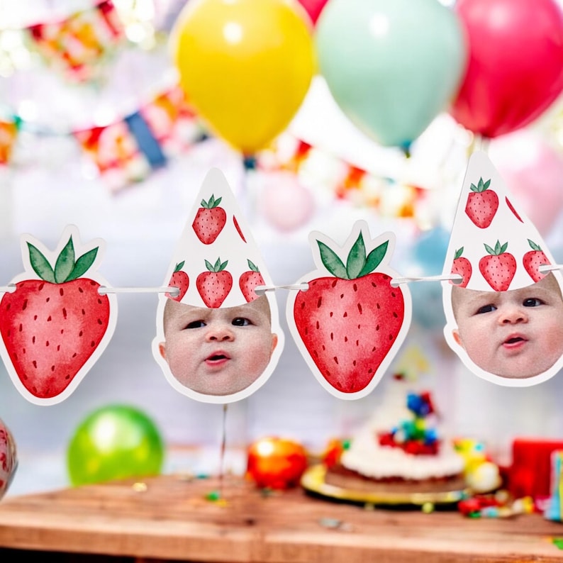 May include: A white banner with red and green strawberries and baby faces. The banner is strung with white string and has a party hat on each strawberry.