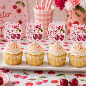 Op de afbeelding: Vier cupcakes op een wit rechthoekig bord, elk voorzien van een papieren prikker met twee rode discobal-kersen, een roze strik en de tekst She's the Cherry on top. De cupcakes zijn voorzien van witte glazuur met roze spikkels.