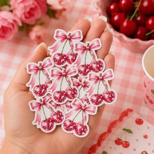 Op de afbeelding: Een verzameling gestanste stickers met paren roze discobal-kersen bekroond met lichtroze linten. Elke sticker toont twee bolvormige, gefacetteerde kersvormen met een reflecterende, glinsterende textuur, verbonden door steeltjes aan een strik.